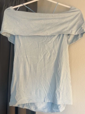 Ljusblå offshoulder topp - Säljer en ljusblå offshoulder topp med bred krage och ärmlös design. Toppen har en avslappnad passform och tunna axelband som ger en trendig look. Perfekt för dig som gillar stilrena och enkla plagg.