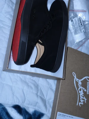 Svarta Louis Junior Flat Velours - Säljer ett par svarta Louis Junior Flat Velours sneakers från Christian Louboutin. Skorna har en klassisk låg siluett, svart mocka och den ikoniska röda sulan. Perfekta för dig som vill ha en clean och lyxig look med streetvibe. Storlek 46 går att diskutera pris