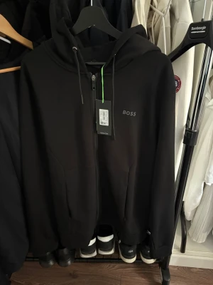 Svart hoodie med dragkedja från BOSS - Snygg svart hoodie från BOSS med dragkedja framtill och justerbar huva med snören. Diskret BOSS-logga på bröstet och två fickor framtill. Perfekt för en clean och stilren look. Tillverkad i mjukt material för extra komfort. Den passar xl-xxl