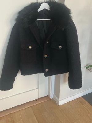 Svart kappa med fuskpäls - Svart kappa med stor fluffig fuskpäls runt kragen och dekorativa metallknappar framtill. Två stora fickor med lock och boxig, trendig passform. Perfekt för dig som vill ha en varm och snygg jacka med attityd. Originalpris 800