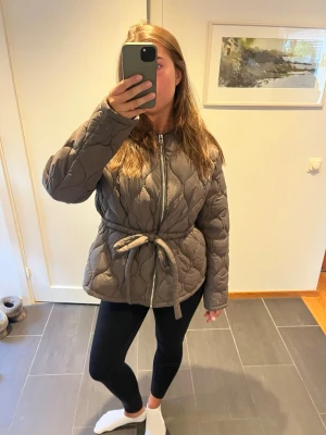 Brun quiltad pufferjacka med knytskärp - Säljer en brun pufferjacka med quiltat mönster och knytskärp i midjan. Jackan har dragkedja framtill och en lite längre, avslappnad passform. Perfekt för kalla dagar och riktigt snygg till både jeans och leggings.