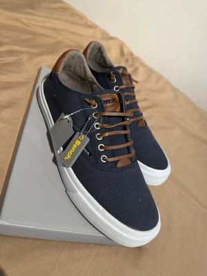 Mörkblå sneakers från Bench - Snygga mörkblå sneakers från Bench med vita sulor och bruna detaljer i läderimitation på plös, häl och snörning. Ovandelen är i textil och skorna har klassisk låg profil. Perfekta för en stilren och avslappnad look.