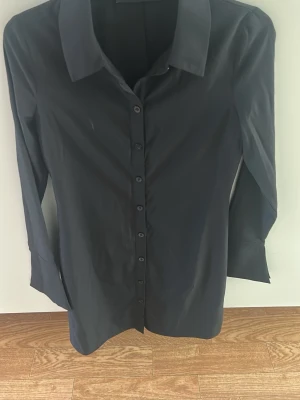 Svart skjortklänning med lång ärm - En svart skjortklänning med klassisk krage och knappar framtill. Klänningen har långa ärmar med breda manschetter och rak passform. Perfekt för en clean och stilren look.