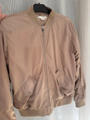 Beige bomberjacka med fickor - Stilren beige bomberjacka med dragkedja framtill och två snedställda fickor med knapp. Jackan har ribbade muddar vid ärmslut, nederkant och krage. Tillverkad i ett glansigt material som ger en modern look.