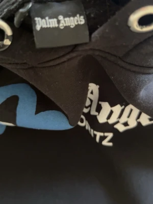 Palm Angels svart hoodie St. Moritz - Säljer en svart hoodie från Palm Angels med trycket 'St. Moritz' framtill. Hoodien har en stor huva med dragsko och coolt blått och vitt tryck i graffiti-stil på bröstet. Perfekt för dig som gillar streetwear och vill sticka ut.