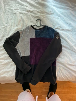 La Vichy tröja i svart och lila - Unik långärmad tröja med patchwork-design i svart, grått, mörkblått och lila. Tröjan har rund halsringning och en liten broderad detalj på ena ärmen. Perfekt för dig som gillar en kreativ och färgstark stil.