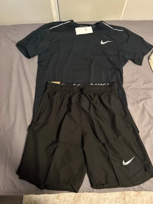 Svart Nike träningsset t-shirt & shorts - Säljer ett svart träningsset från Nike med t-shirt och shorts. Båda plaggen har den klassiska Nike-loggan i vitt. T-shirten har rund hals och korta ärmar, shortsen har resår i midjan och sidofickor. Perfekt för gymmet eller löpning.