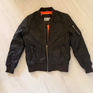 Svart bomberjacka Urban Classics - Snygg svart bomberjacka från Urban Classics med orange innerfoder. Klassisk modell med ribbade muddar, dragkedja framtill och fickor på sidorna samt en ärmficka med dragkedja. Perfekt för en streetwear-look. Har inte använt den alls mycket så tycker den är i jättefint skick. 