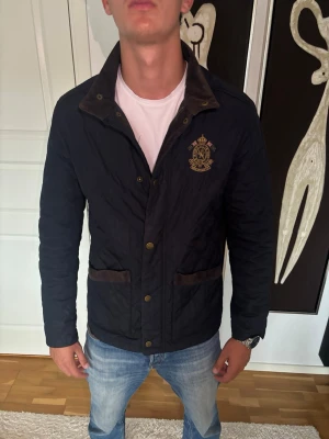 Fieldjacket - Snygg mörkblå quiltad jacka med brunt detaljer och ett broderat emblem på bröstet. Jackan har knappar framtill, två fickor och långärmad design. Perfekt för dig som vill ha en stilren och klassisk look.