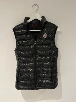 Svart dunväst från Moncler - Snygg svart dunväst från Moncler med glansig finish och quiltad design. Västen har hög krage, dragkedja framtill och Moncler-logga på bröstet. Perfekt för lager-på-lager och håller dig varm under kyliga dagar. Storlek xs för tjejer