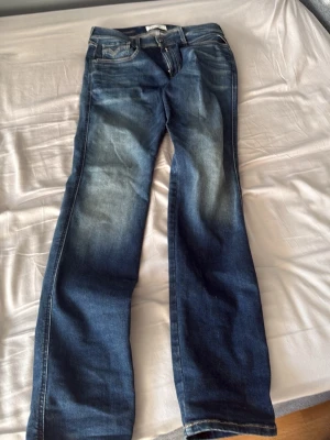 Blå jeans från Replay, modell Anbass hyperflex - Säljer ett par blå jeans från Replay, modell Anbass. Jeansen har en klassisk femficksdesign, raka ben och snygga slitningar framtill. Tillverkade i denim med kontrastsömmar och metallknapp i midjan. Perfekta för en avslappnad och trendig look.
