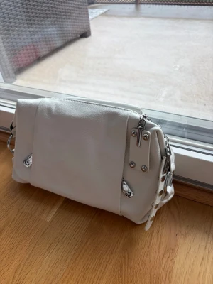Vit handväska med silverdetaljer - Snygg vit handväska i mjukt material med flera silverfärgade dragkedjor och nitar. Väskan har en modern, lite boxig form och praktiska fack både fram och bak. Perfekt accessoar för att lyfta din outfit.
