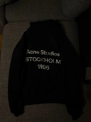 Svart hoodie från Acne Studios - Snygg svart hoodie från Acne Studios med tryck på ryggen där det står 'Acne Studios STOCKHOLM 1996'. Klassisk passform med huva och ribbade muddar. Perfekt för dig som gillar stilrena och trendiga plagg.