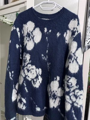 Mörkblå stickad tröja med blommor - Snygg mörkblå stickad tröja från ASOS DESIGN med stora vita blommor över hela tröjan. Tröjan har rund halsringning och långa ärmar. Perfekt för dig som gillar en trendig och mysig look.
