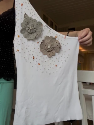 Vit oneshoulder topp med blommor och strass  - Snygg vit oneshoulder topp med två stora dekorativa blommor i beige och små pärlor och stenar framtill. Toppens material känns mjukt och stretchigt, perfekt för en trendig look. Passar dig som vill sticka ut med coola detaljer och asymmetrisk design. Märke : Papareil