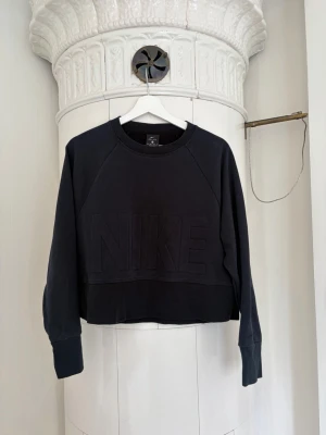 Svart croppad Nike sweatshirt - Svart croppad sweatshirt från Nike med stor NIKE-logga i relief på bröstet. Tillverkad i Dri-FIT-material som andas och har breda muddar vid ärmsluten. Perfekt för en sportig och chill look.