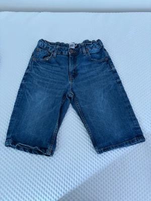 Blå jeansshorts med resårmidja - Säljer ett par klassiska blå jeansshorts med resår i midjan och femficksmodell. Shortsen har normal passform och är tillverkade i slitstark denim, perfekta för sommardagar. Passar dig som gillar en enkel och tidlös stil.