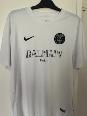 Vit PSG x Balmain fotbollströja - Snygg vit fotbollströja med PSG-emblem och Balmain Paris-tryck på bröstet. Tröjan har rund hals, korta ärmar och är tillverkad i ett lätt, andningsbart material som passar perfekt till träning eller match. Nike-logga i svart på framsidan.