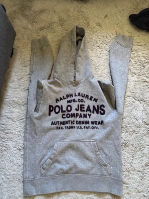 Sällsynt Grå hoodie Polo Jeans Ralph Lauren - Snygg grå hoodie från Polo Jeans Company Ralph Lauren med stor svart text på bröstet. Klassisk känguruficka framtill och huva. Perfekt för en avslappnad streetwear-look. Mjuk bomullsblandning och ribbade muddar vid ärmar och midja.