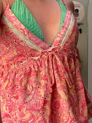 Rosa och gul omlottklänning med spets - Söt kort omlottklänning i rosa och gult med paisley-inspirerat mönster. Klänningen har spetsdetaljer vid urringningen, knyt i midjan och tunna axelband. Perfekt för varma dagar och ger en bohemisk vibe.