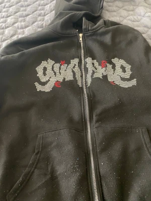 Svart hoodie med blingad text - Säljer en svart hoodie med dragkedja och huva. På bröstet finns ett stort tryck i blingade stenar som bildar ett coolt textmotiv med små röda detaljer. Hoodien har fickor framtill och en avslappnad passform, perfekt för en streetwear-look.