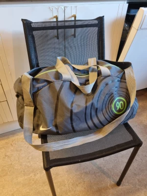 Nike Total 90 Duffelväska / Sportväska - Säljer en grå duffelväska från Nike med ljusgråa handtag och axelrem. Väskan har ett coolt cirkelmönster i svart och grönt samt en grön rand på dragkedjan. Perfekt för resor eller tennis, badminton, gymmet, rymmer mycket och är lätt att bära.