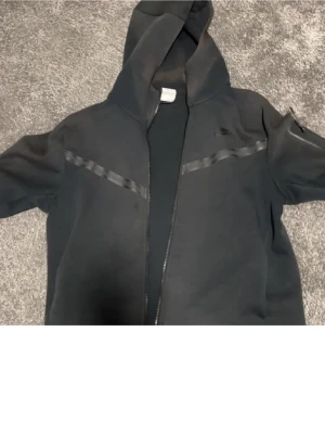 Svart nike tech fleece - Säljer en svart hoodie med dragkedja och huva. Hoodien har diskreta svarta detaljer i form av band över bröstet och ärmarna. Perfekt för en avslappnad och stilren look. Tillverkad i mjukt material för extra komfort.