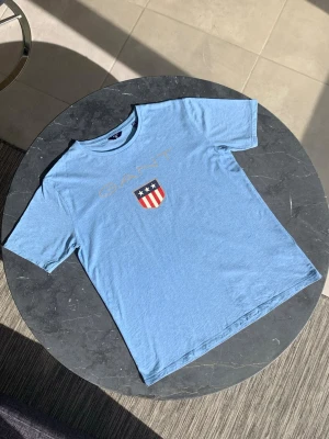 Blå GANT t-shirt med tryck NY! - Snygg ljusblå t-shirt i nyskick från GANT med stort tryck framtill i form av logga och sköld med stjärnor och ränder. Klassisk rund hals och korta ärmar. Perfekt till jeans eller shorts för en avslappnad look.