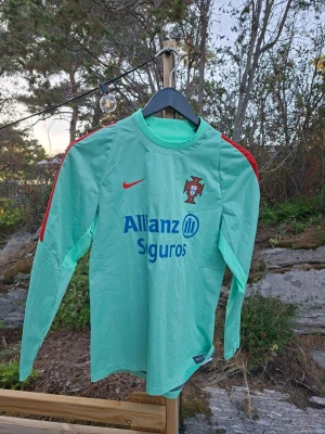  Portugal långärmad träningströja - Snygg långärmad träningströja i mintgrönt med röda detaljer på axlarna och Nike-logga. Portugal-emblem på bröstet och sponsortryck från Allianz Seguros. Perfekt för fotbollsträning eller chill dagar. Tillverkad i lätt och funktionellt material. 