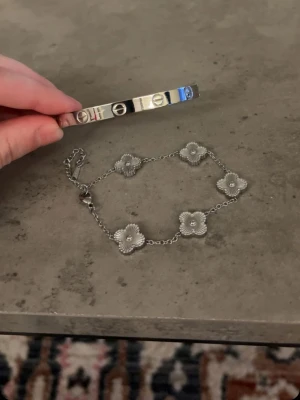 Silverfärgade armband med blommor - Två stilrena armband i silverfärg. Det ena är ett stelt armband och det andra är ett kedjearmband med fem klöver i metall, där varje blomma har en liten pärldetalj i mitten. Perfekta accessoarer för att lyfta din outfit. ‼️HELT NYA ALDRIG ANVÄND och det är STAINLESS STEEL alltså tappar ej färg‼️