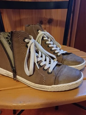 Bruna sneakers från Rieker med snörning - Säljer ett par bruna sneakers från Rieker med vita snören och dragkedja på sidan. Skorna har en vit sula och är tillverkade i mocka. Insidan har ett blommigt mönster. Perfekta för dig som gillar en avslappnad och stilren look.