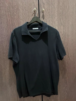 Svart polotopp från Zara - Klassisk svart polotopp från Zara med korta ärmar och mjukt material. Toppen har en bred krage och enkel, stilren design som passar till det mesta. Perfekt för dig som gillar minimalistisk stil.