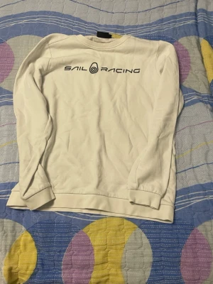 Vit långärmad tröja Sail Racing - Vit långärmad tröja från Sail Racing med tryckt logga framtill och en liten logga i nacken. Klassisk rund halsringning och ribbade muddar vid ärmslut och nederkant. Perfekt till en avslappnad och stilren look.