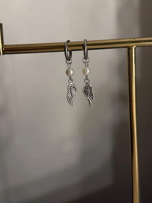 Silvriga örhängen med pärlor och vingar - Säljer ett par silvriga örhängen med små vita pärlor och hängande vingeformade berlocker. Örhängena har en enkel och stilren design som passar perfekt till många olika looks. Materialet ser ut att vara metall och pärlorna ger en extra touch.