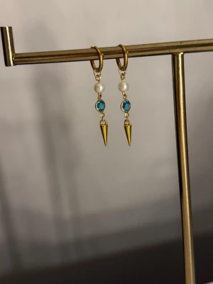 Guldörhängen med pärlor och blå sten - Snygga örhängen i guldton med små vita pärlor, blå glittrande stenar och spetsiga hängen. Perfekta för att ge din look en cool och elegant touch. Hängande modell med trendig design.