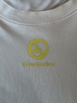 Vit Acne Studios t-shirt med tryck - Vit t-shirt, "Acne Studios" med gult tryck och logga på bröstet. Hade på mig den en gång men den va lite större än jag tänkte mig. Det är storlek S, oversized. Passar även M. Priset är lågt för en anledning 👀