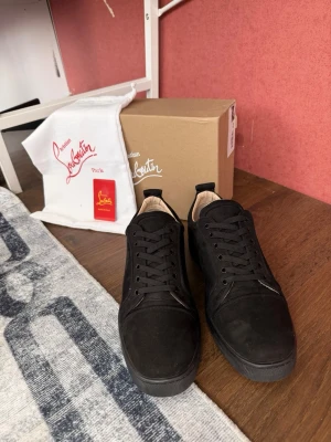 Svarta sneakers från Christian Louboutin - Stilrena svarta sneakers från Christian Louboutin i mocka med ton-i-ton detaljer och klassisk låg siluett. Skorna har svarta skosnören, diskret logga broderad på hälen och en svart yttersula. Perfekta för dig som gillar en clean och exklusiv look.