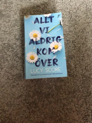 Allt vi aldrig kom över - En romantisk roman av Lucy Score med blått omslag och prästkragar. Boken handlar om Naomi som flyttar till småstaden Knockemout i Virginia för att hjälpa sin tvillingsyster och möter frisören Knox. Perfekt för dig som gillar romantik, känslor och spänning.
