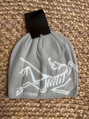 Grey Arc’teryx beanie - Snygg ljusgrå mössa från Arc'teryx med vitt skelettmönster på framsidan. Tillverkad i mjukt stickat material som sitter skönt på huvudet. Perfekt för kalla dagar och enkel att matcha med olika outfits.