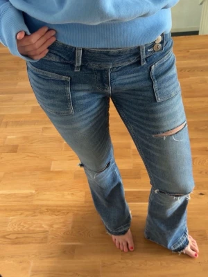 Blå slitna bootcut jeans - Säljer ett par blå bootcut jeans med slitningar och hål på knäna. Jeansen har två framfickor, dubbla knappar i midjan och en klassisk tvättad look. Perfekta för en avslappnad och trendig stil. Storlek M men passar även S