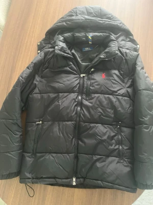Svart pufferjacka från Polo Ralph Lauren - Svart pufferjacka med huva från Polo Ralph Lauren. Jackan har dragkedja framtill, två sidofickor med dragkedja och en liten röd logga på bröstet. Klassisk quiltad design som håller värmen under kalla dagar.