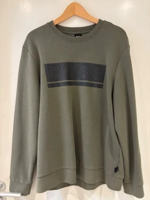 Olivgrön sweatshirt från HUGO BOSS - Snygg olivgrön sweatshirt från BOSS med stort svart logotryck på bröstet. Tröjan har rund halsringning, långa ärmar och ribbade muddar vid ärmslut och nederkant. Perfekt för en avslappnad och stilren look.