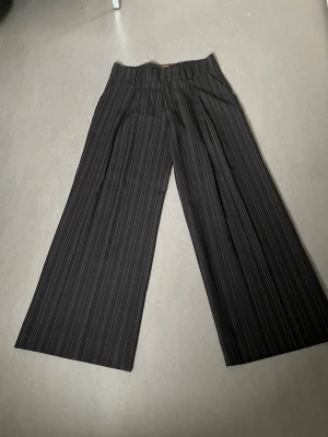 Svarta kostymbyxor med vita ränder - Snygga svarta kostymbyxor med tunna vita kritstrecksränder och hög midja. Byxorna har vida ben och pressveck framtill för en klassisk look. Perfekta för dig som gillar en stilren och trendig outfit.