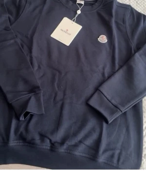 Mörkblå sweatshirt från Moncler - Snygg mörkblå sweatshirt från Moncler med rund halsringning och diskret Moncler-logga på bröstet. Tröjan har långa ärmar och ribbade muddar vid ärmslut och nederkant. Perfekt för en clean och stilren look.