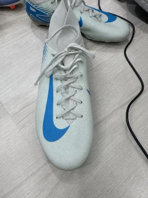 Nike Mercurial Vapor dobbar  - Vita fotbollsskor från Nike med stor blå Swoosh på sidan och blå grafik vid hälen. Skorna har snörning och är gjorda i ett lätt syntetmaterial som ger bra känsla på planen. Perfekta för dig som vill ha stilrena och funktionella skor. Pris kan diskuteras.