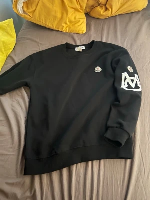 Svart hoodie med tryck på ärmen pris kan diskuteras  - Svart hoodie med rund hals och ribbade muddar. Har ett litet broderat märke på bröstet och ett större vitt tryck på ena ärmen. Perfekt för en stilren och avslappnad look.
