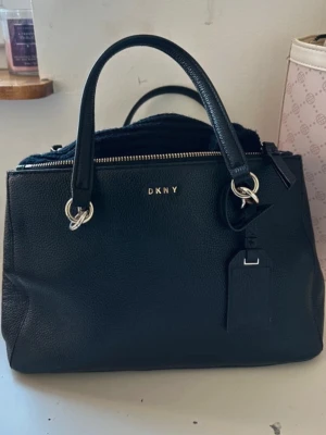 Svart handväska från DKNY i skinn - Snygg svart handväska från DKNY i  skinn med silverfärgade metalldetaljer. Har aldrig använt den