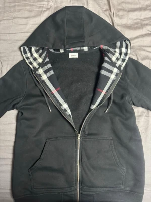 Svart hoodie med rutigt foder från burberry - Säljer en svart hoodie med dragkedja och klassiskt rutigt foder i huvan. Hoodien har fickor framtill, dragsko i huvan och ett diskret broderat motiv på bröstet. Perfekt för dig som gillar stilrena detaljer och bekväma plagg.