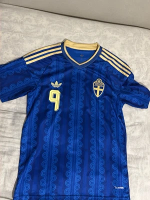 Blå Sverige fotbollströja Adidas - Säljer en blå Sverige fotbollströja från Adidas med gula detaljer, nummer 9 och namnet ISAK på ryggen. Tröjan har mönstrad design, korta ärmar och klassiska Adidas-ränder på axlarna. Perfekt för dig som vill stötta landslaget med stil.