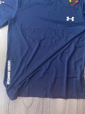 Blå träningströja från Under Armour - Snygg blå träningströja från Under Armour med logga på bröstet och tryck längs sidan. Tillverkad i ett lätt och snabbtorkande funktionsmaterial som passar perfekt till gymmet eller löprundan. Klassisk rund hals och korta ärmar.